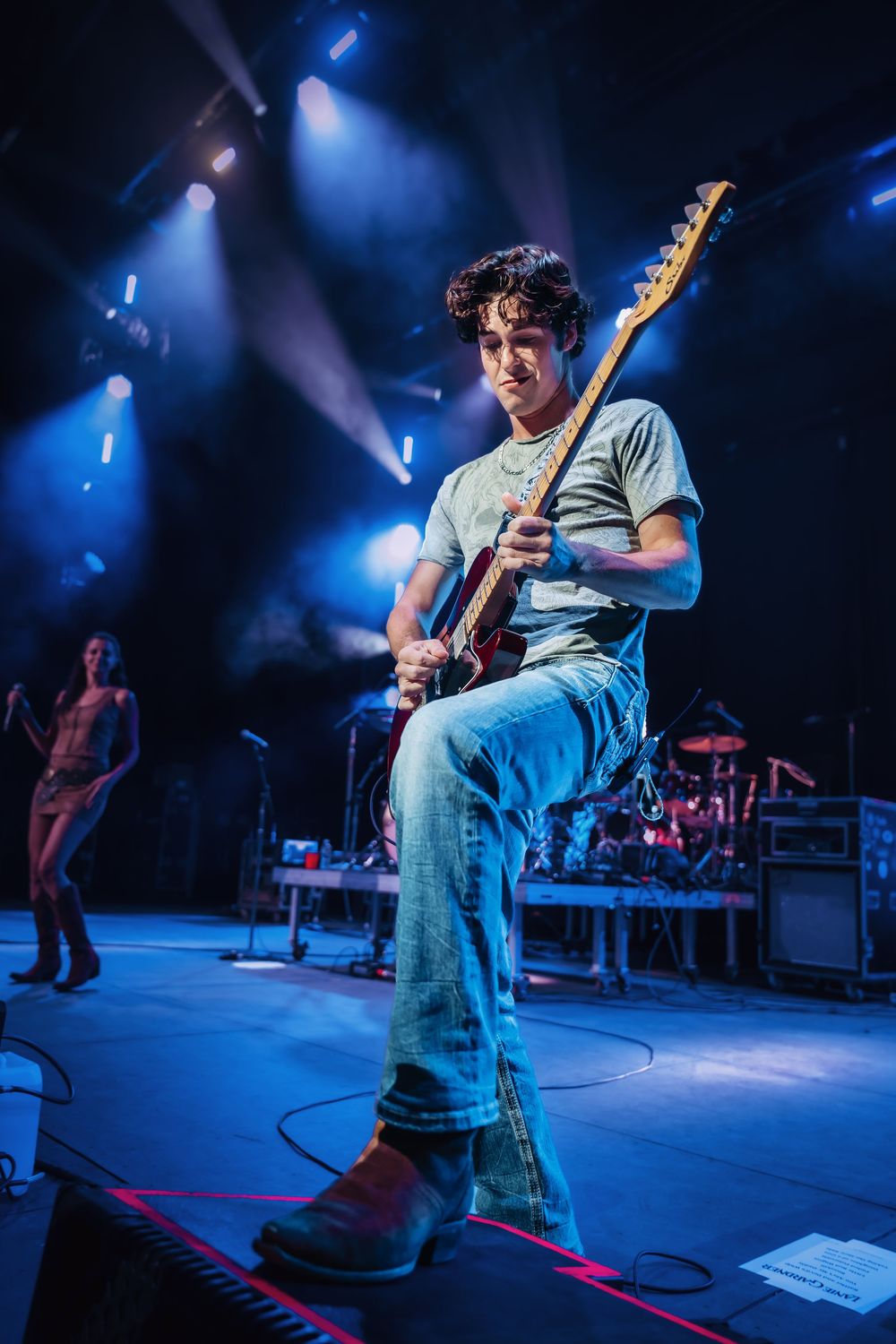 Young male guitarist performing on stage with blue concert lighting and band members visible in background.