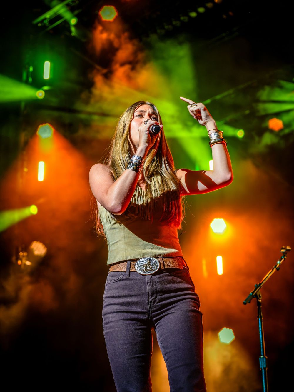 Female singer performing on stage with microphone, colorful green and orange stage lights illuminating smoke behind her.
