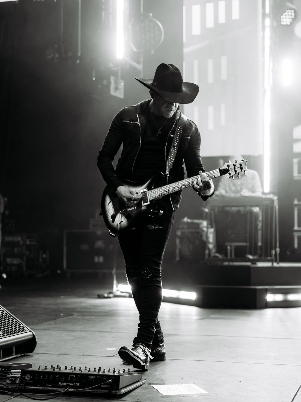 Black and white photo of a guitarist in cowboy hat and leather jacket performing on a lit concert stage.