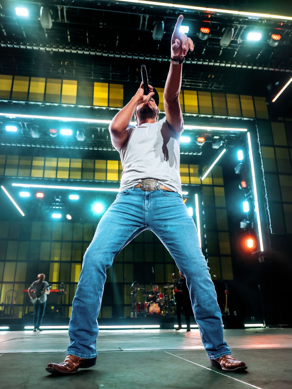 Male performer in white tank top and jeans raises hand on stage with colorful lights behind him.