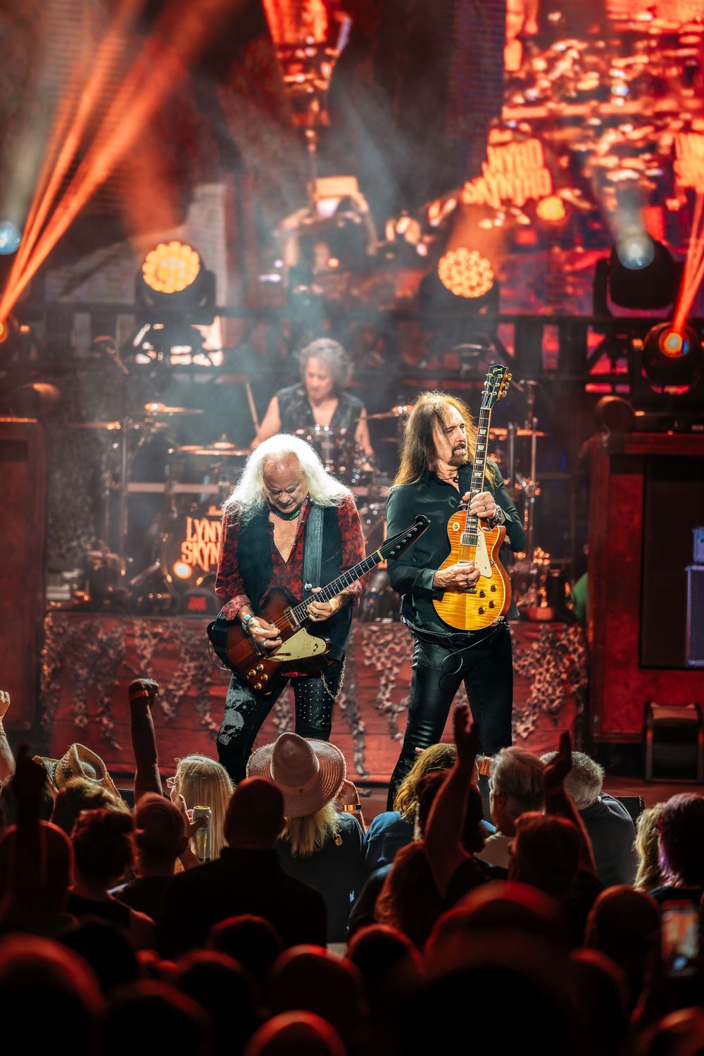 Two guitarists perform on a dramatic red-lit rock concert stage with crowd in foreground and drummer visible behind.