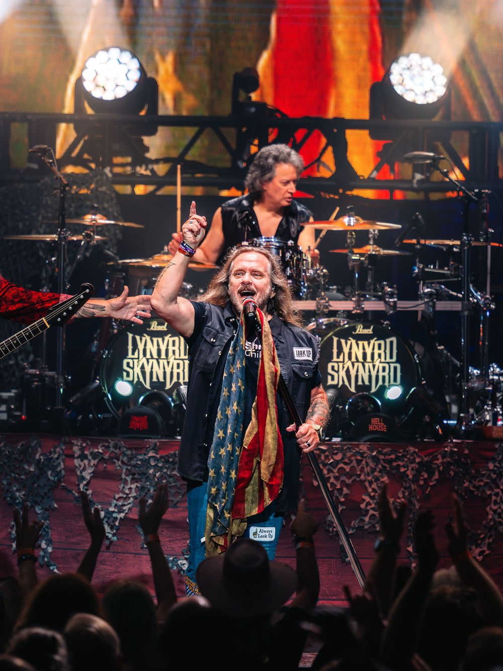 Lynyrd Skynyrd band performing live on stage with lead singer in American flag vest and drummer in background.