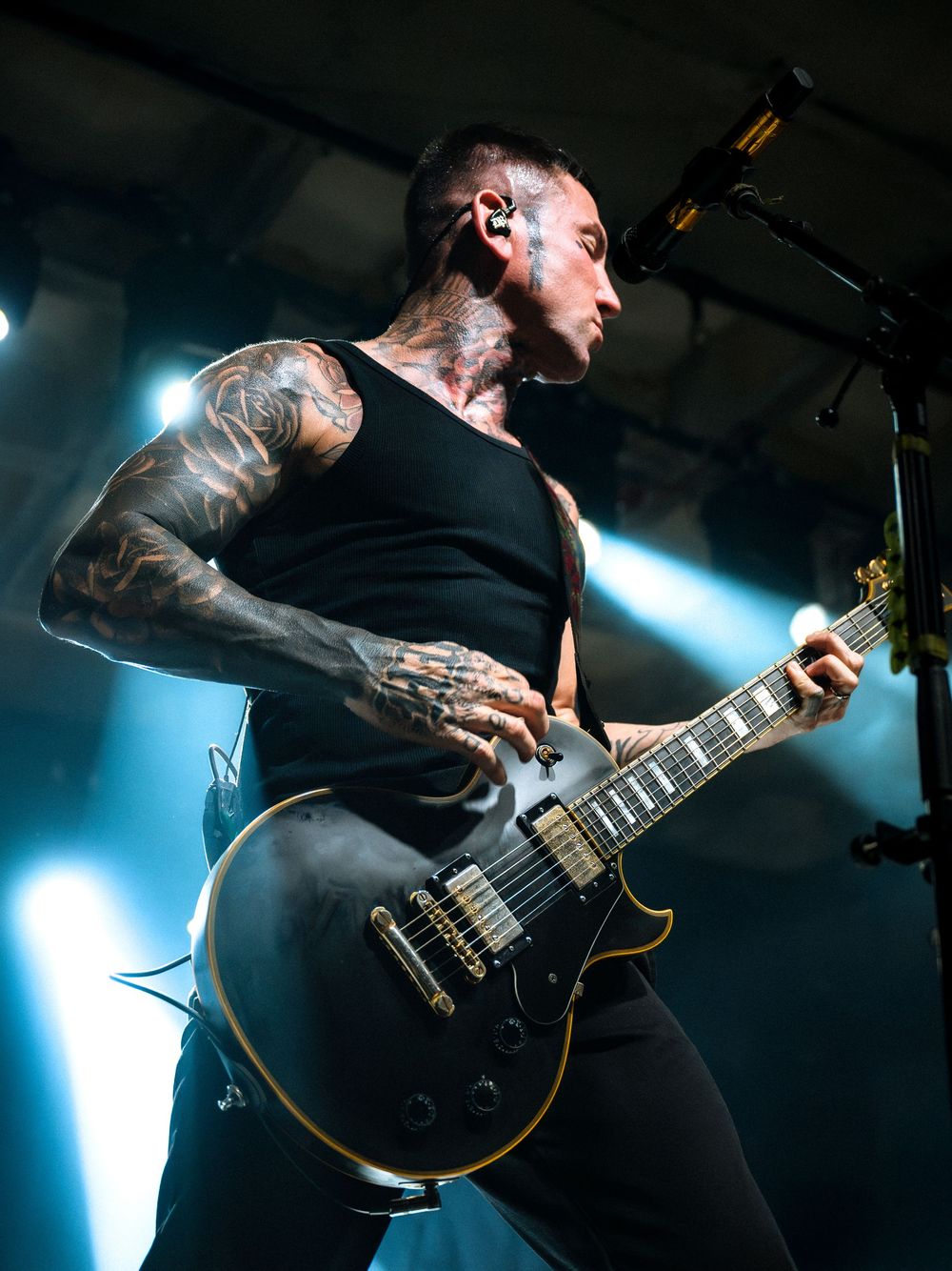 Tattooed male guitarist performing on stage in black tank top, playing dark Les Paul electric guitar under blue stage lights.