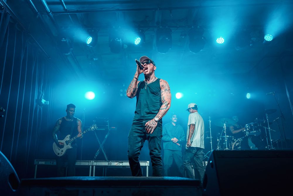 Band performing on stage with blue lighting, lead singer in tank top holding microphone at live concert.