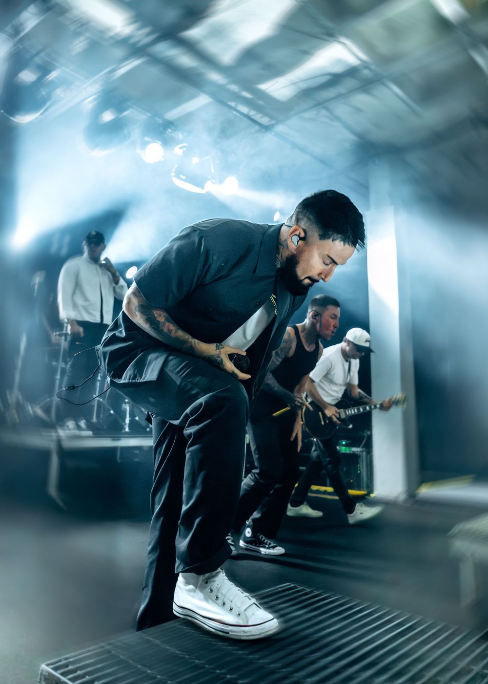 Tattooed performer bends forward on stage under blue lights while band members play guitar in the background.