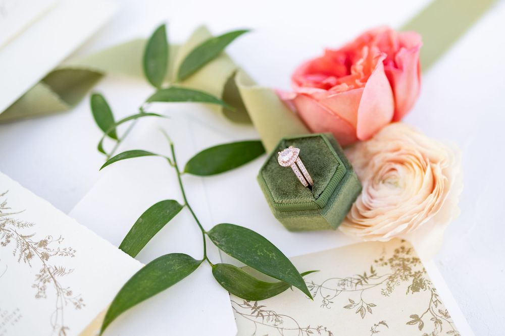 Elegant engagement ring in green velvet box surrounded by pink roses and eucalyptus on a white surface.