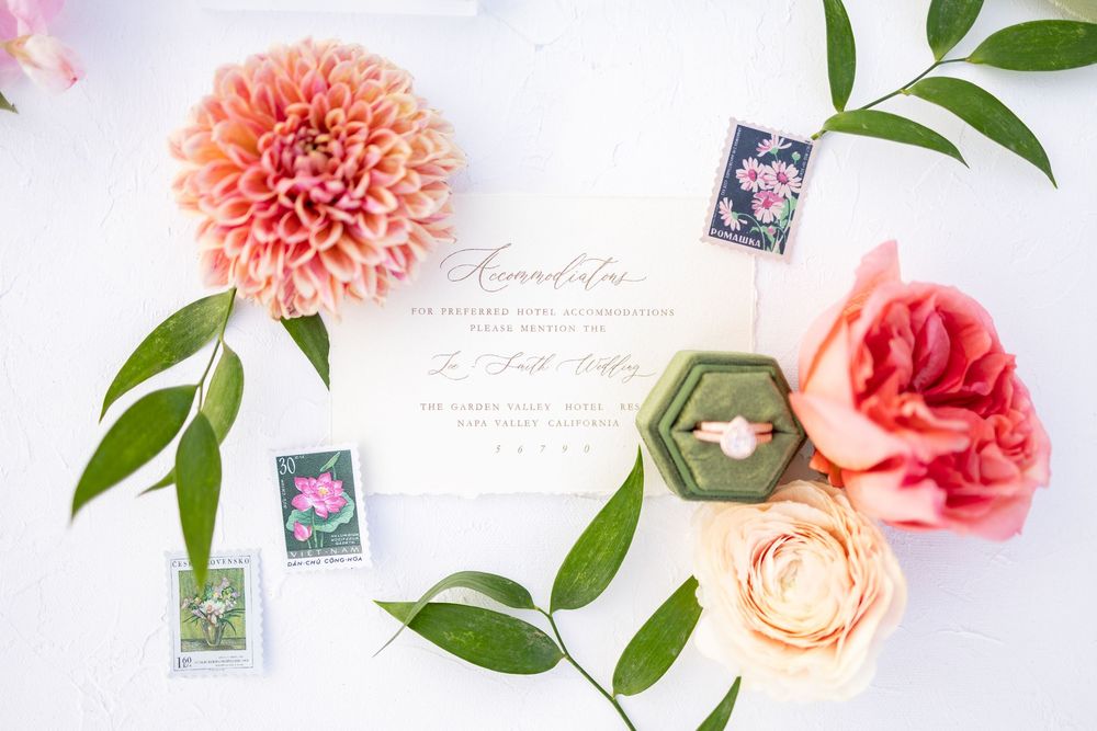 Elegant wedding flat lay with floral invitations, vintage stamps, a ring in a green velvet box, and pink dahlias on white.