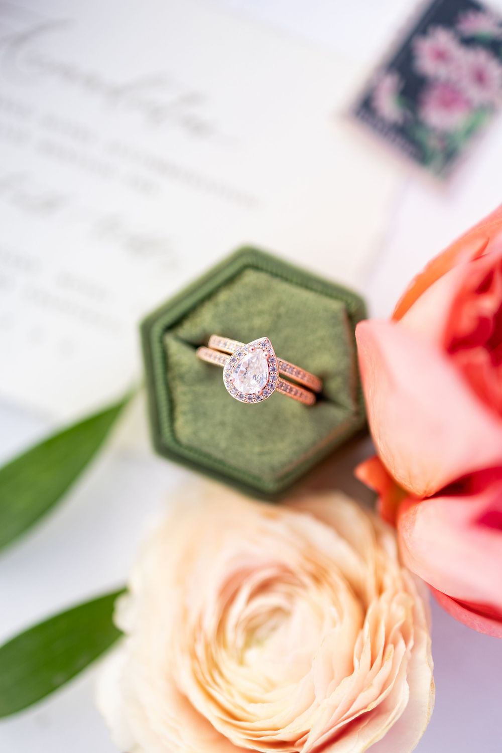 Rose gold pear diamond halo engagement ring with wedding band displayed in green velvet hexagon box surrounded by peach flowers.