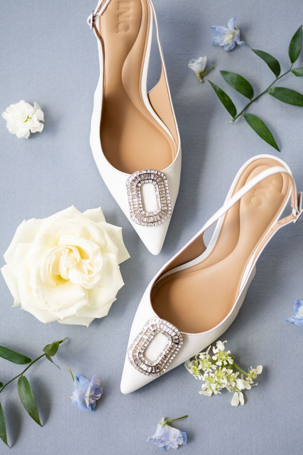 White satin pointed-toe slingback flats with crystal buckle embellishment, surrounded by roses and blue flowers on gray background.