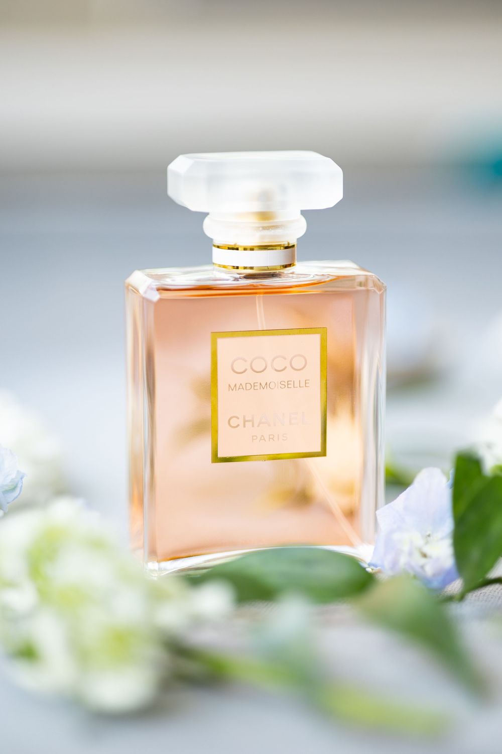 Chanel Coco Mademoiselle perfume bottle surrounded by delicate white flowers on a soft gray surface.