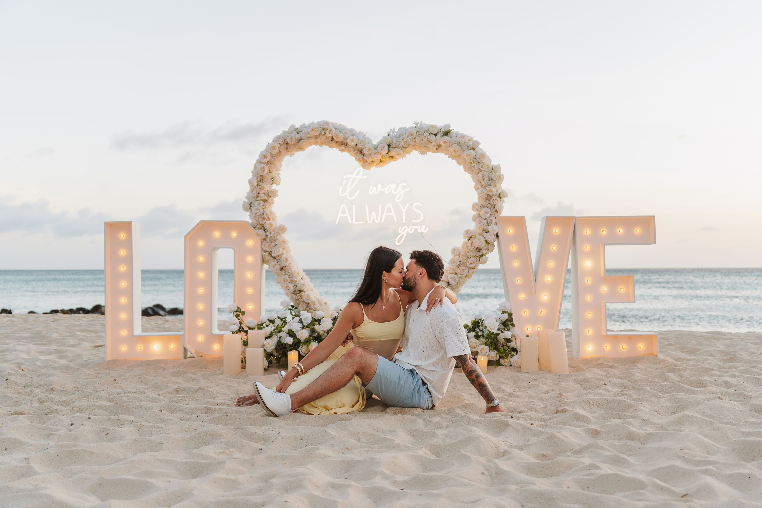 Couple holding hands at sunset beach proposal setup with floral arch, candles, and Marry Me sign on sandy shore.