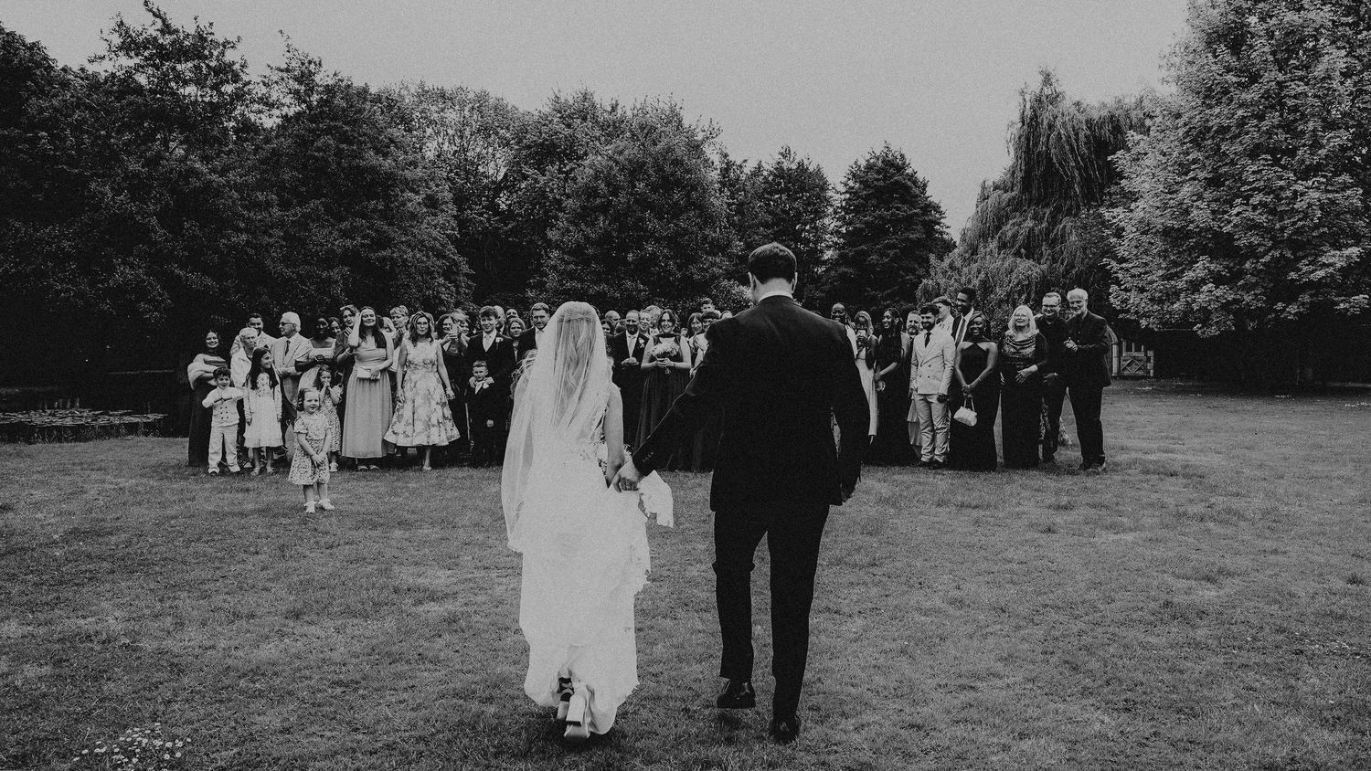 Black and white photo of bride and groom walking toward wedding guests gathered outdoors in a lush garden setting.