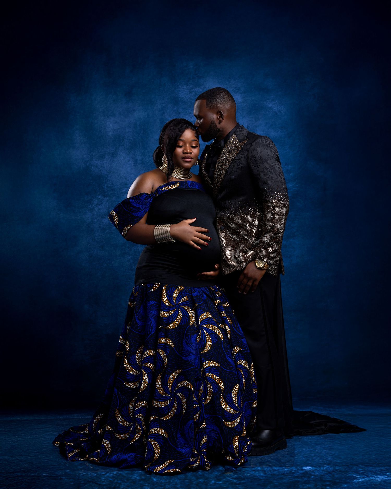 Elegant maternity photo shoot of a couple in blue African print skirt and gold blazer against a dark blue studio backdrop.