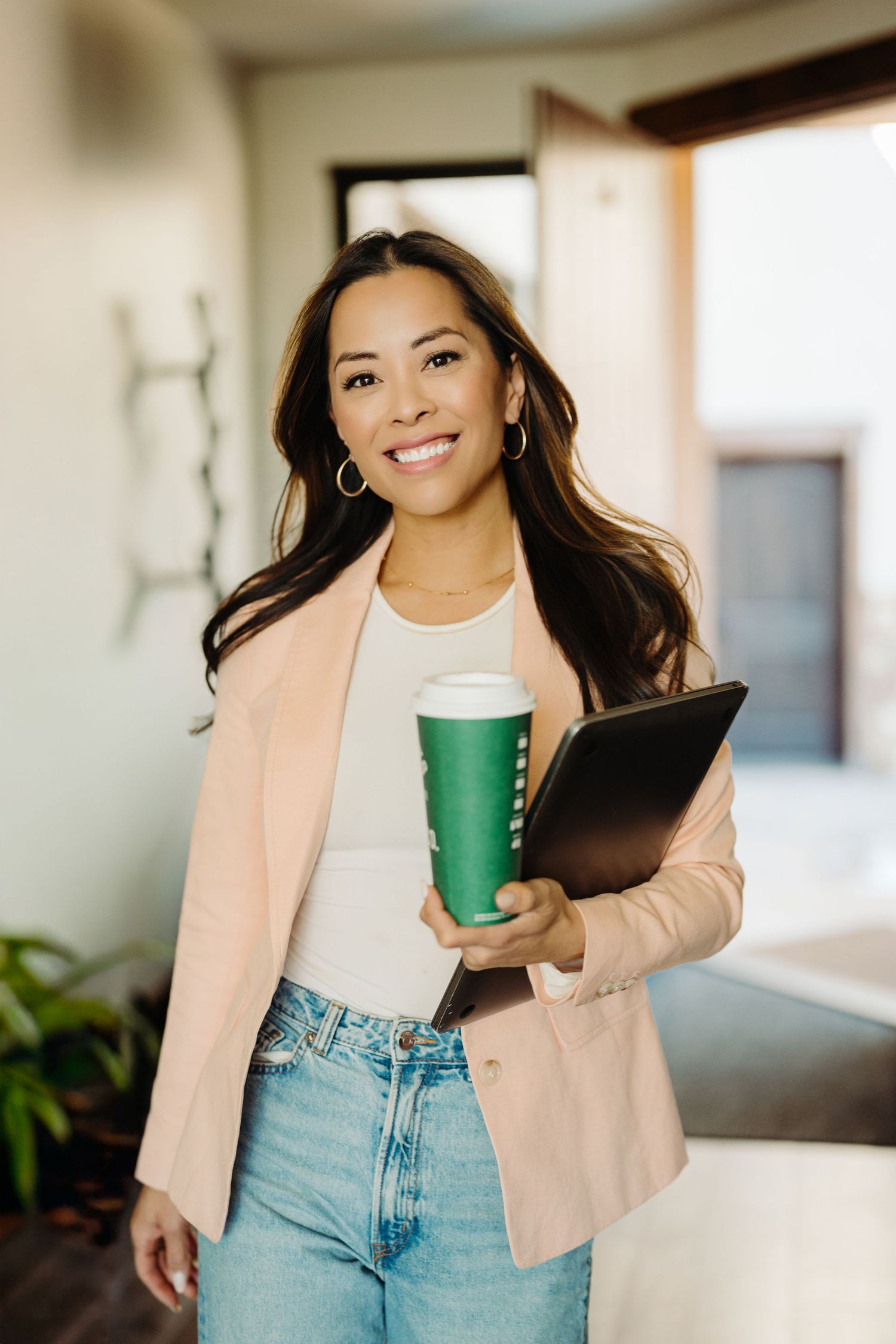 Smiling woman in pink blazer holding green coffee cup and tablet in modern office setting.