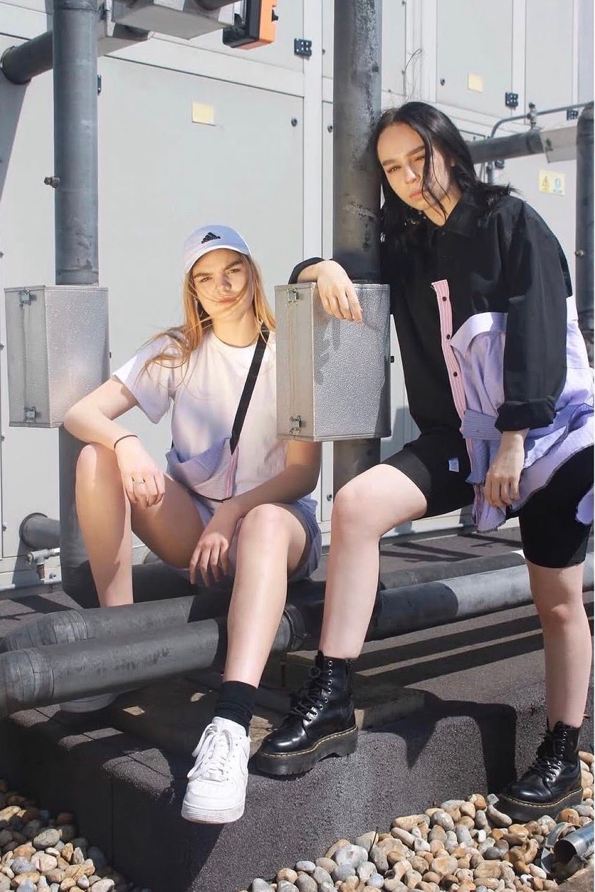 Two young women sitting outdoors in casual streetwear, one in white sneakers, the other in black boots.