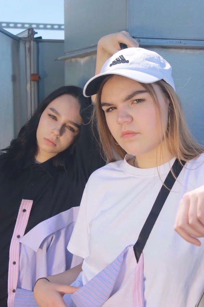 Two young women posing outdoors, one wearing a white Adidas cap and oversized tee, the other in black.