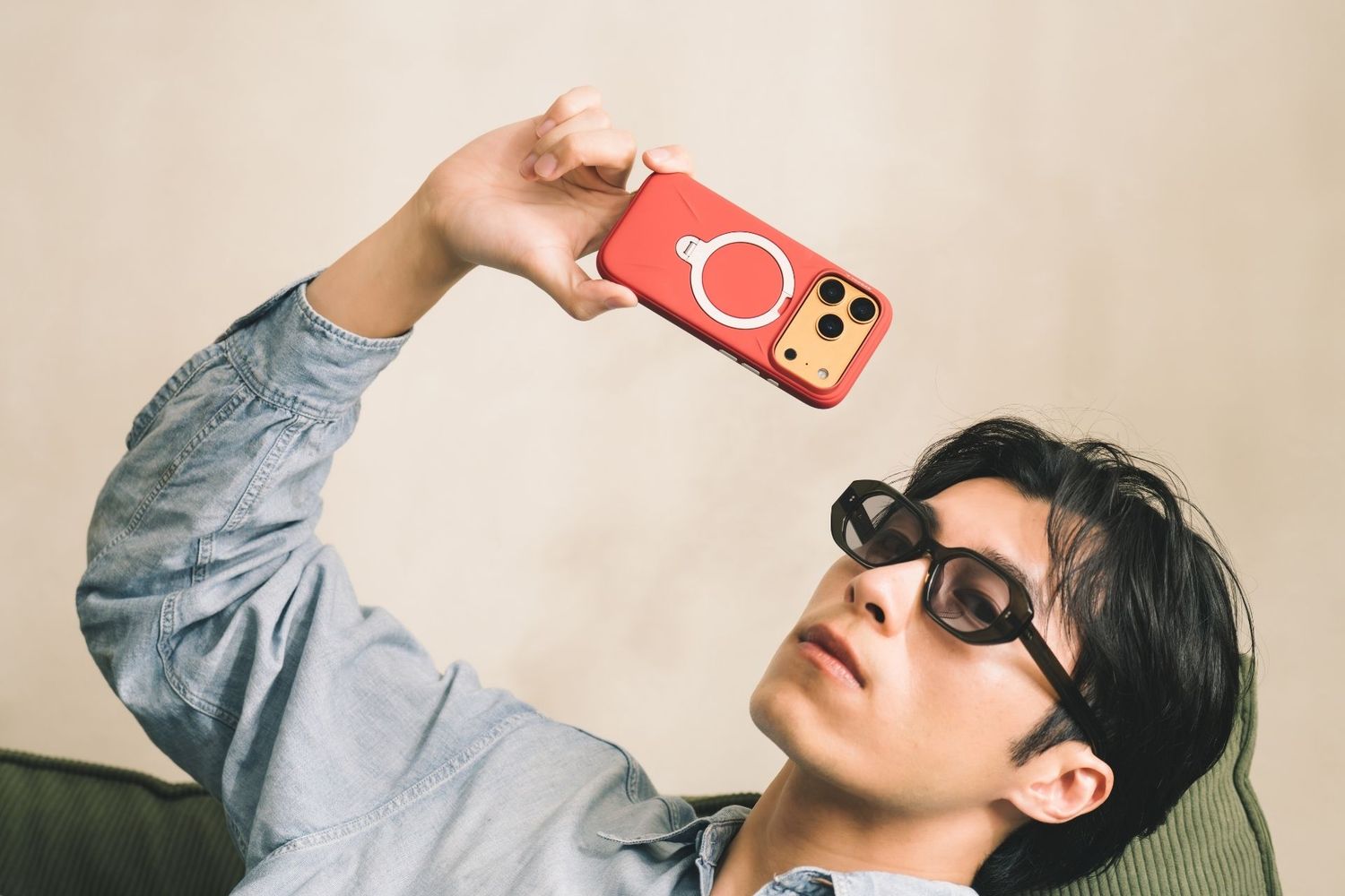 Young man in sunglasses taking a selfie with a red MagSafe iPhone case while reclining on a chair.