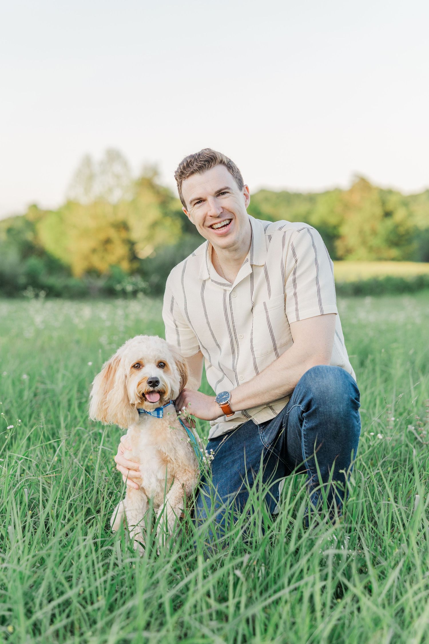 Smiling young man kneeling in green grass field with a fluffy golden doodle dog wearing a blue collar.