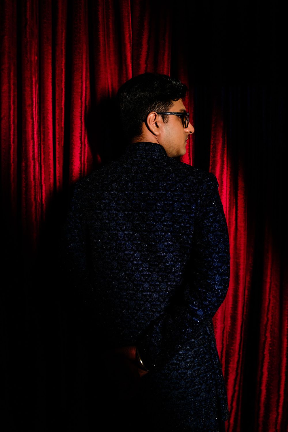Man in dark patterned jacket standing before red velvet curtain, viewed from behind in dramatic lighting.