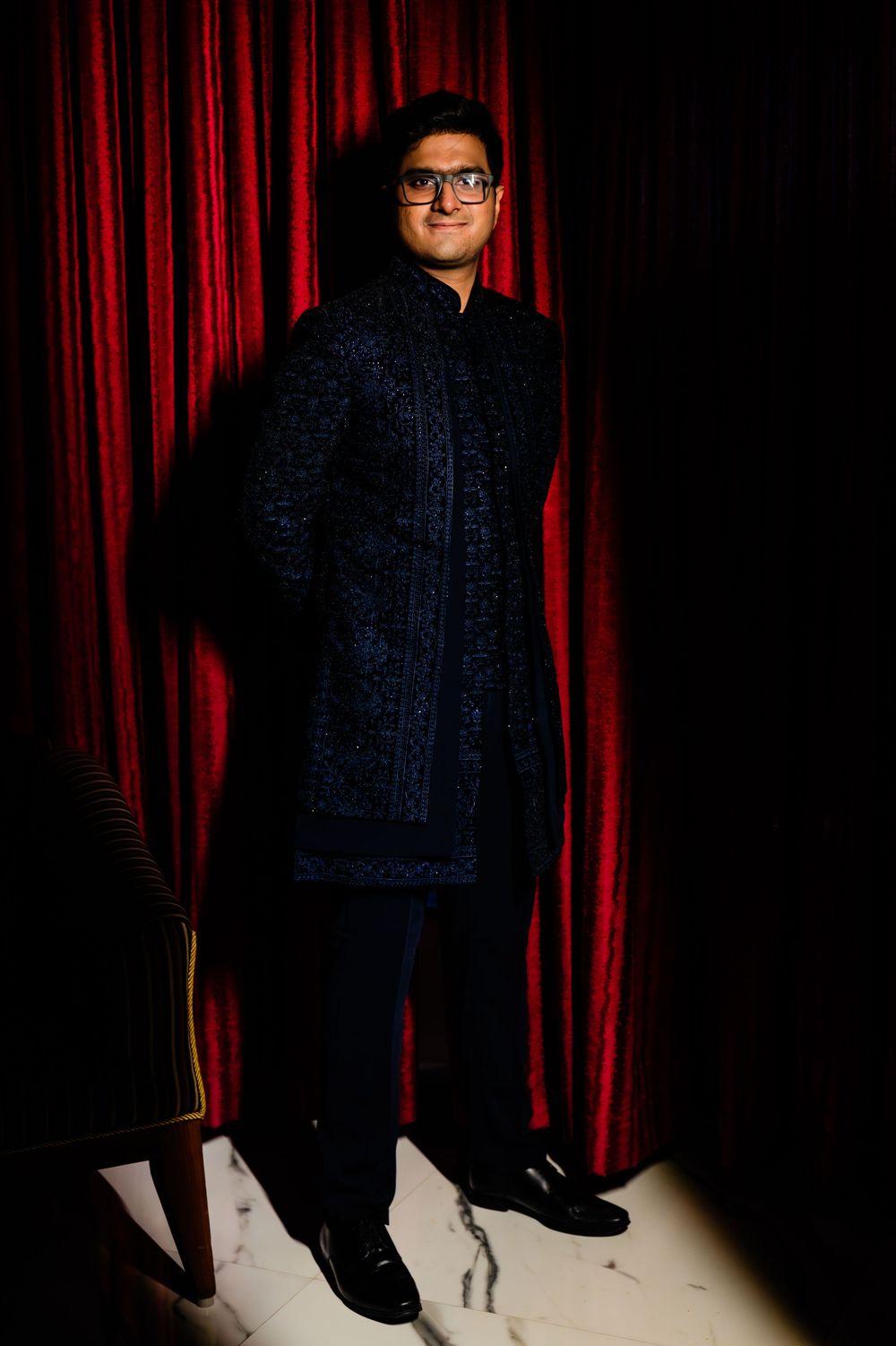 Groom in embroidered navy blue sherwani posing against red curtain backdrop.