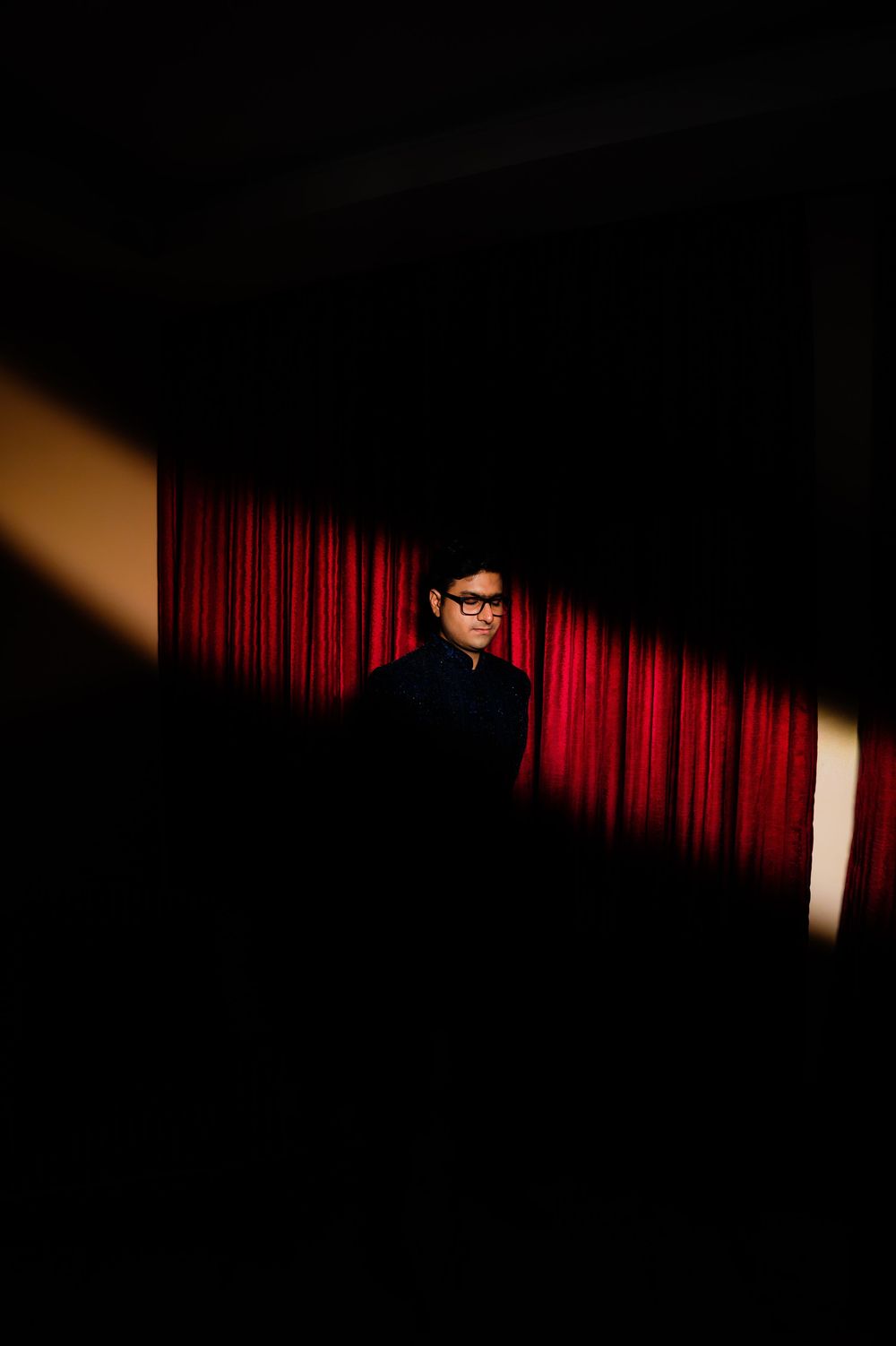 Person in glasses stands before red curtains in dramatic dark lighting, creating a moody theatrical atmosphere.