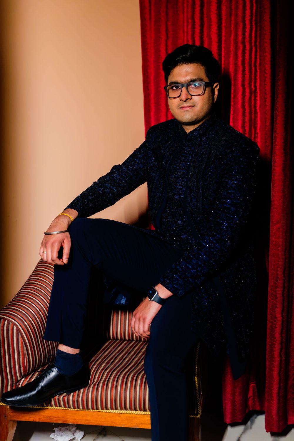 Young man in black embroidered kurta and glasses sitting on striped sofa near red curtain.