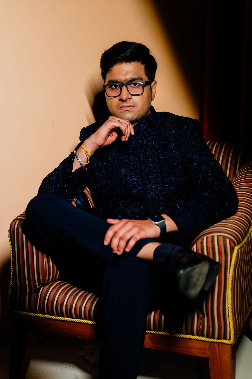 A well-dressed Indian man in a navy embroidered blazer sits confidently in a striped armchair, wearing glasses and a smartwatch.