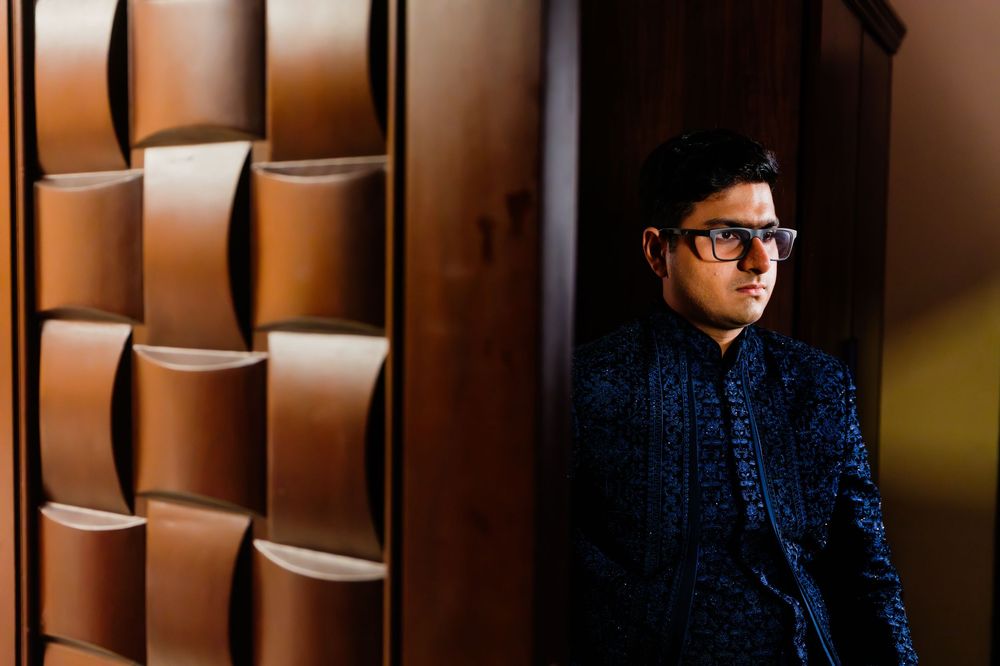 Man in dark embroidered kurta with glasses stands by ornate wooden door in dramatic lighting.