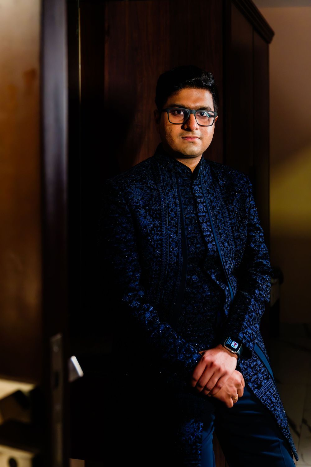 Man in navy blue embroidered sherwani and glasses poses confidently against dark wooden cabinet.