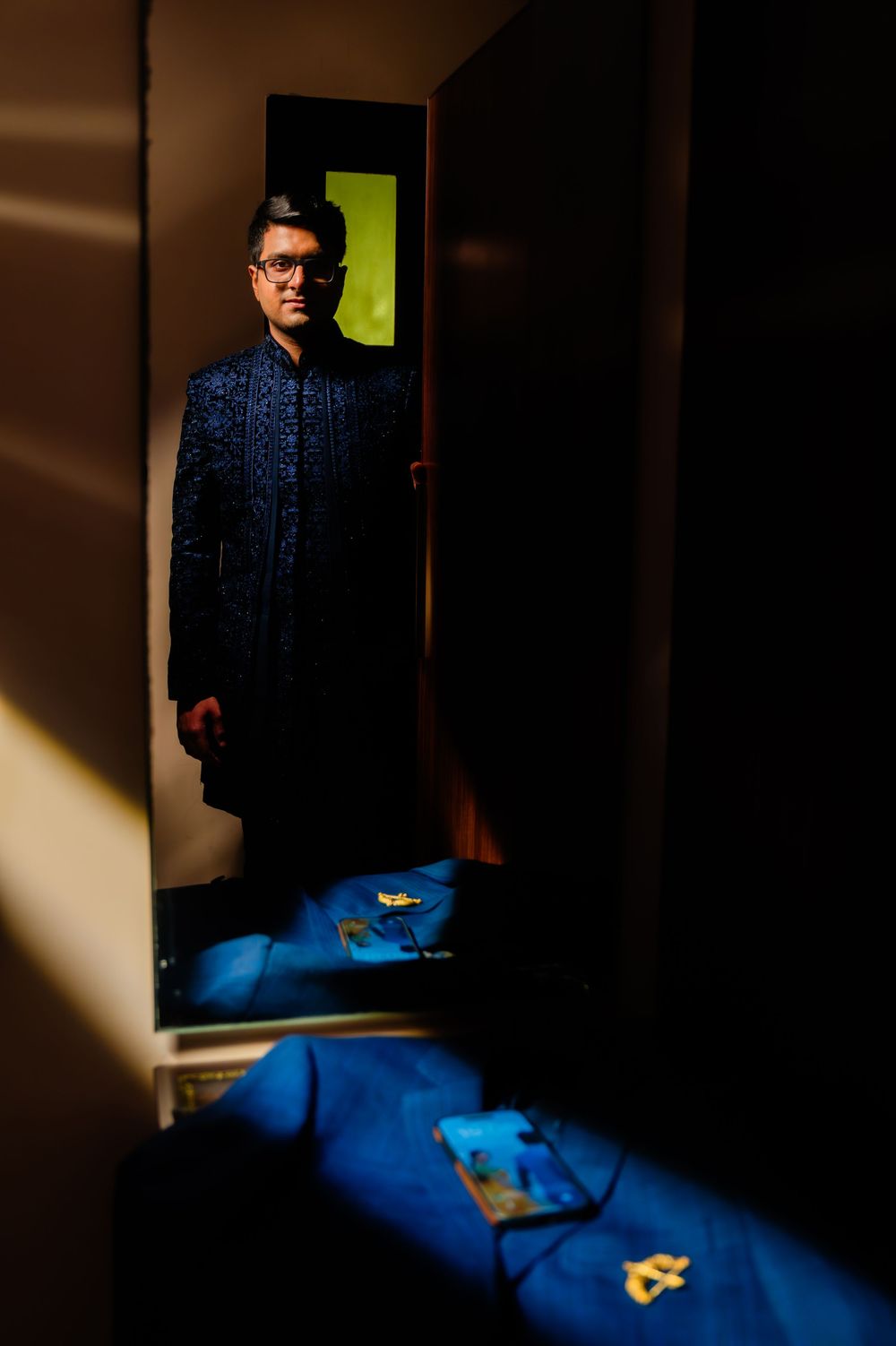 Man in blue embroidered sherwani stands in dramatic doorway with blue fabric and scattered items in foreground.