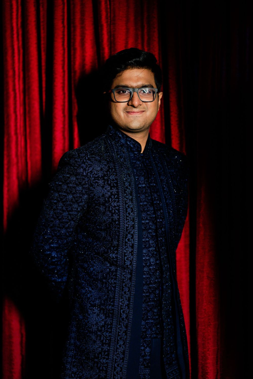 Young man in embroidered navy blue sherwani with glasses posing against a rich red curtain backdrop.