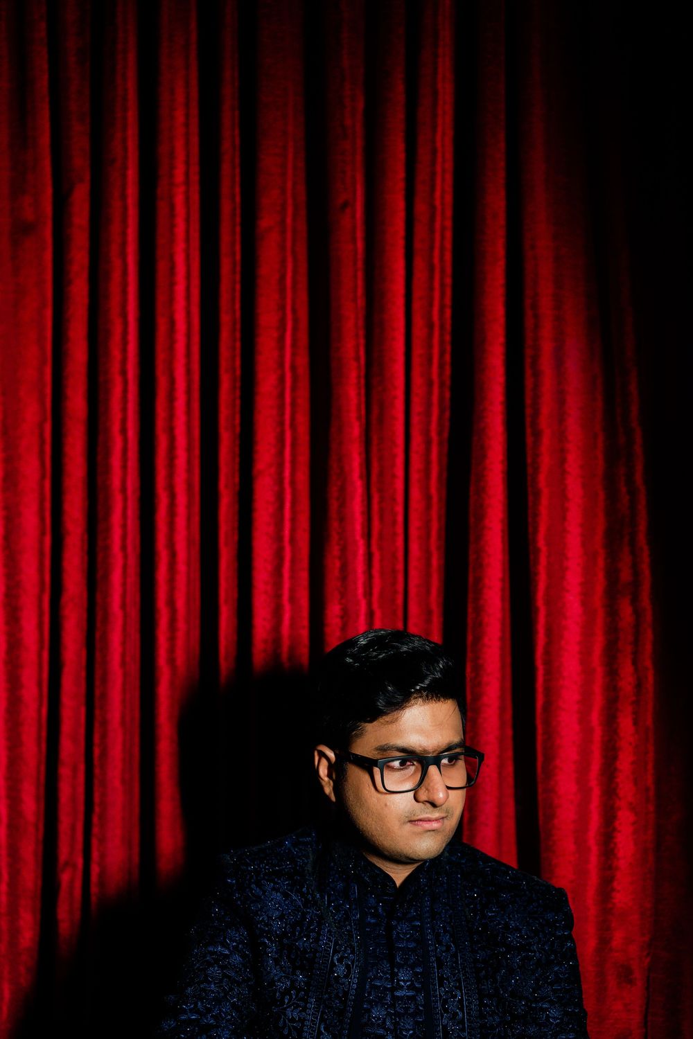 Young man in dark embroidered jacket and glasses poses against rich red velvet curtain backdrop.