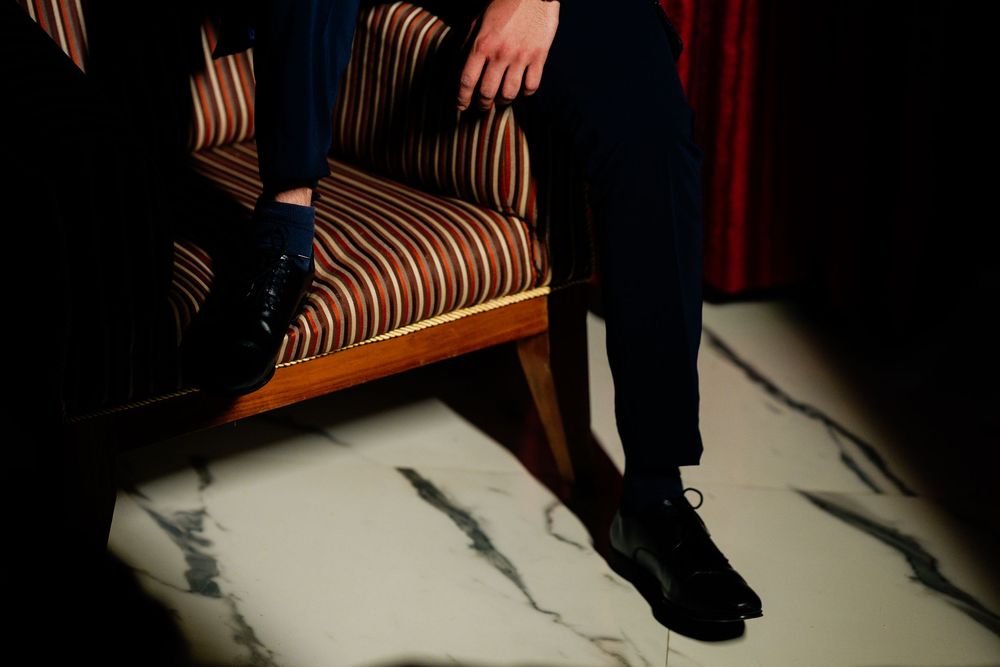 Man in navy trousers and black dress shoes seated on a striped wooden armchair on marble floor.