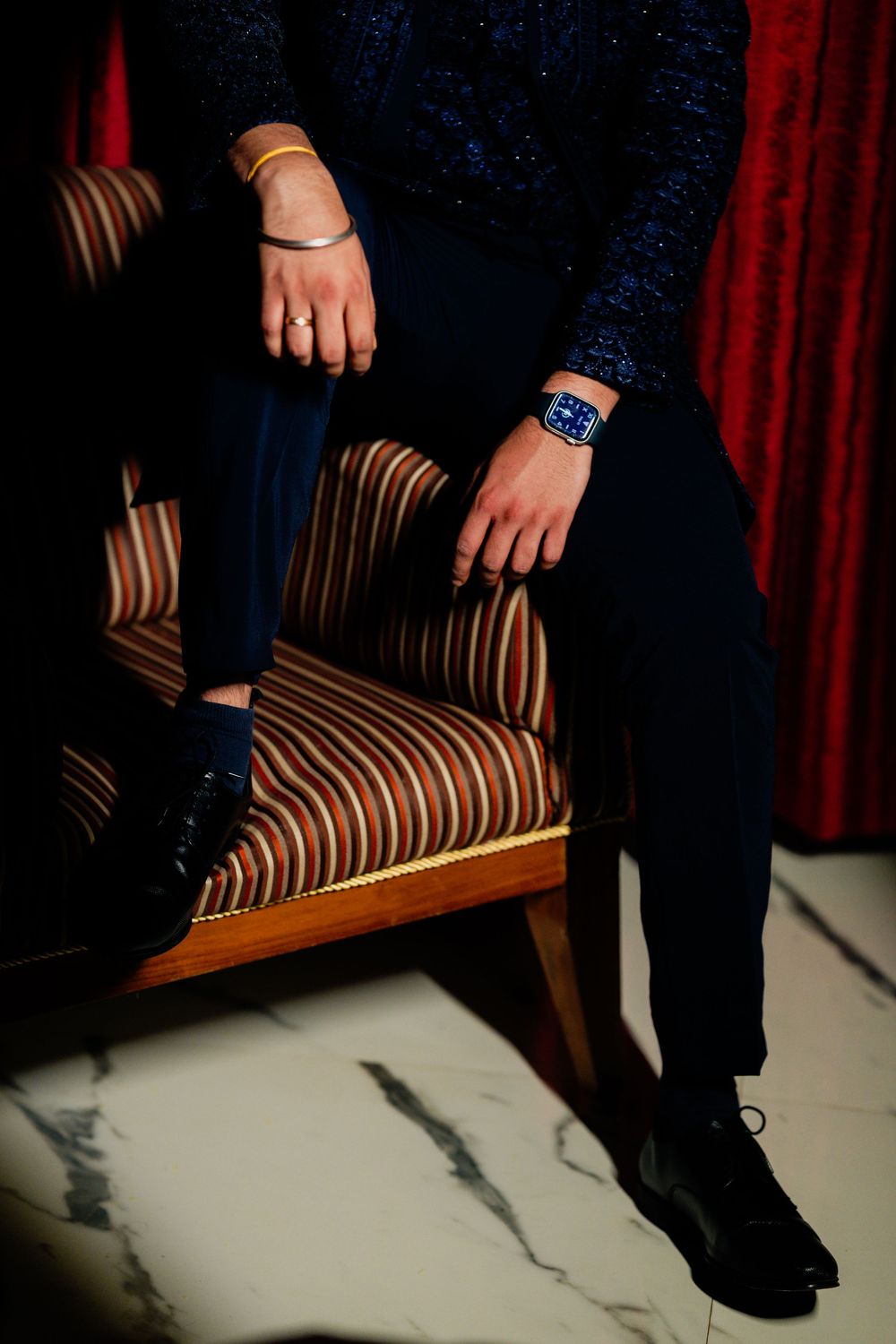Man in navy suit sitting on striped armchair, wearing blue watch and gold bracelet, with black dress shoes.