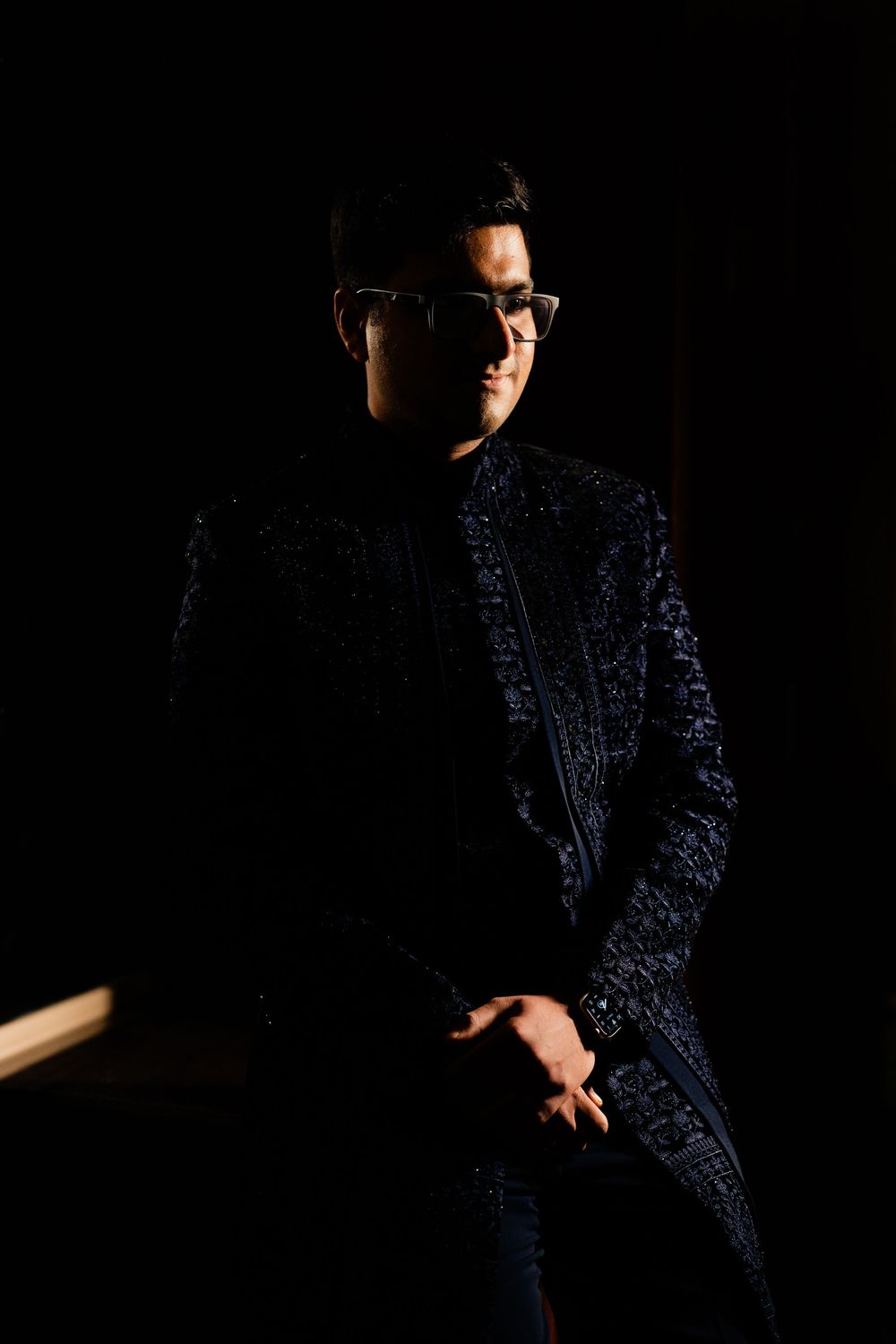 Man in embroidered navy blue suit and glasses posing in dramatic dark studio lighting.
