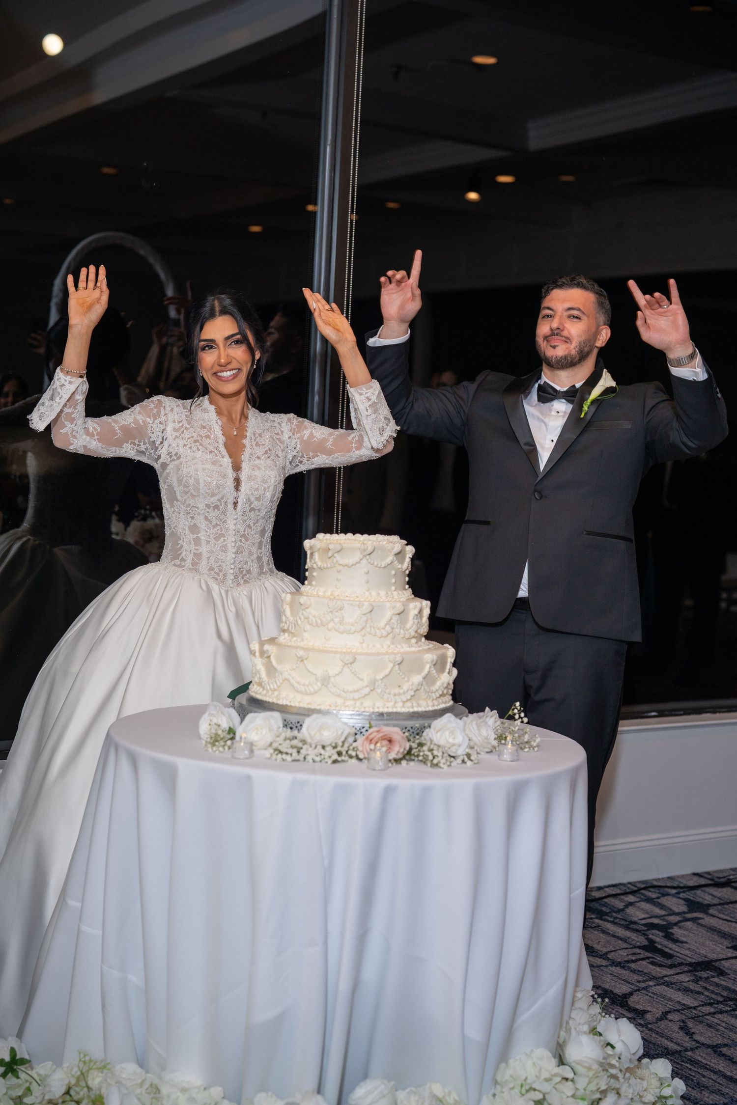 Bride and groom celebrate joyfully beside a white tiered wedding cake at their elegant reception.