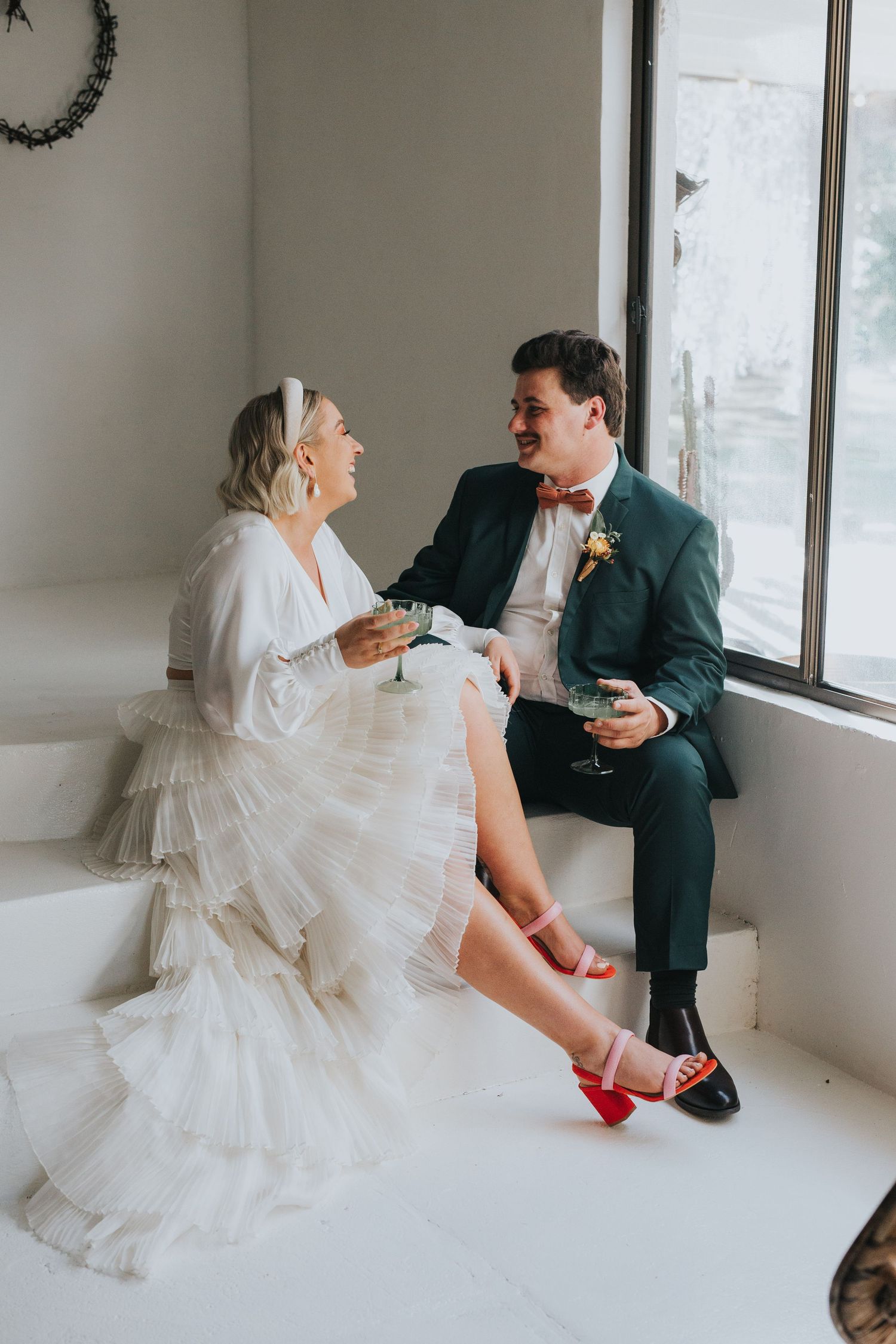 Bride in white ruffled gown and red heels shares a laugh with groom in green suit while sipping cocktails by a window.