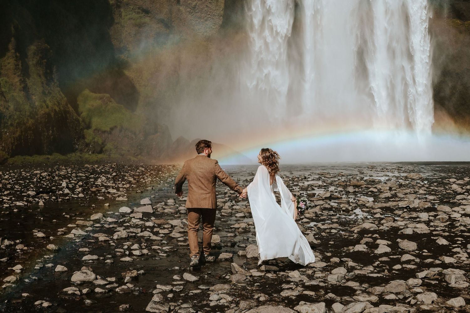 eloping couple running towards Skógafoss waterfall in Iceland while holding hands