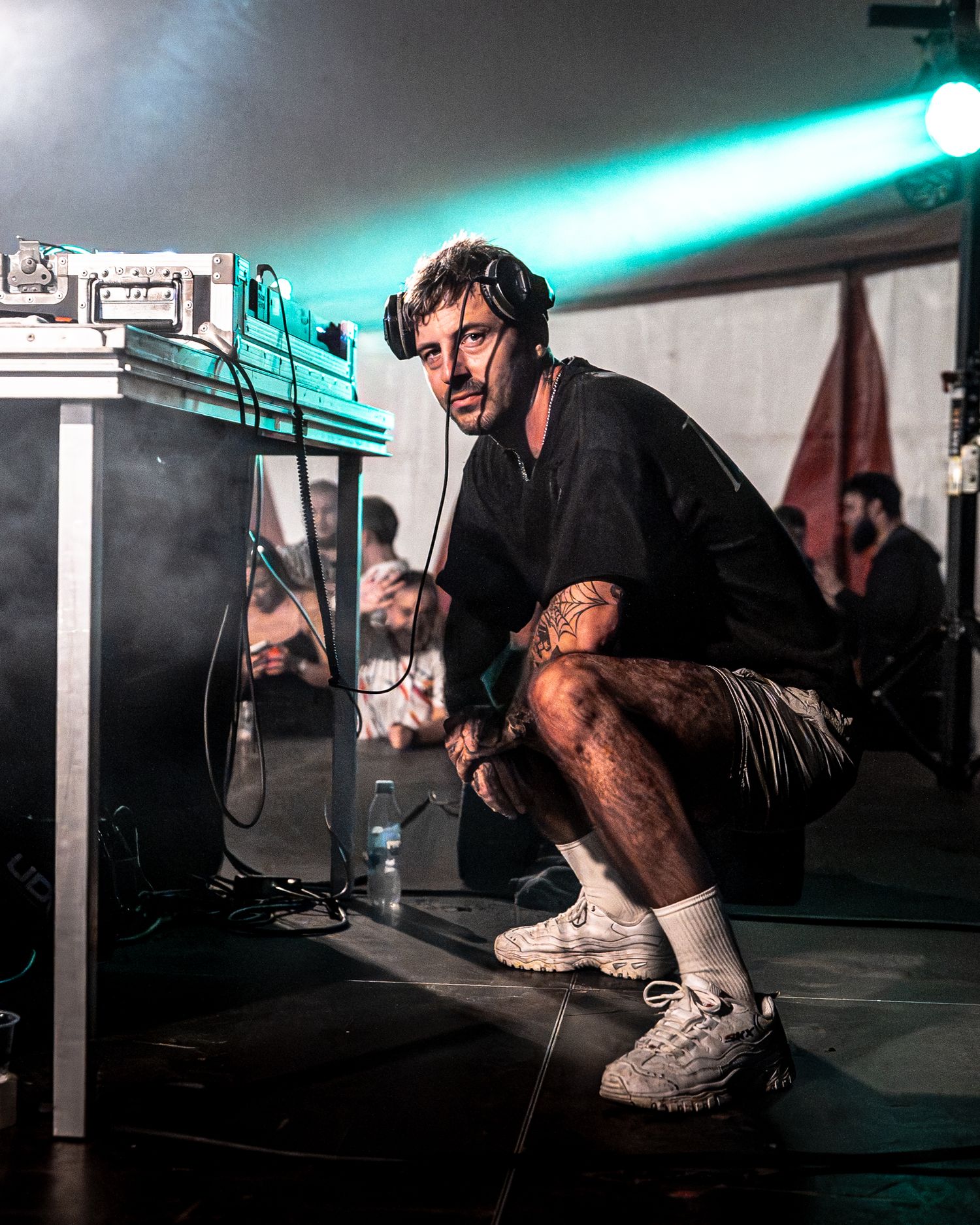 Tattooed male DJ crouching on stage near equipment during live music event with crowd in background.