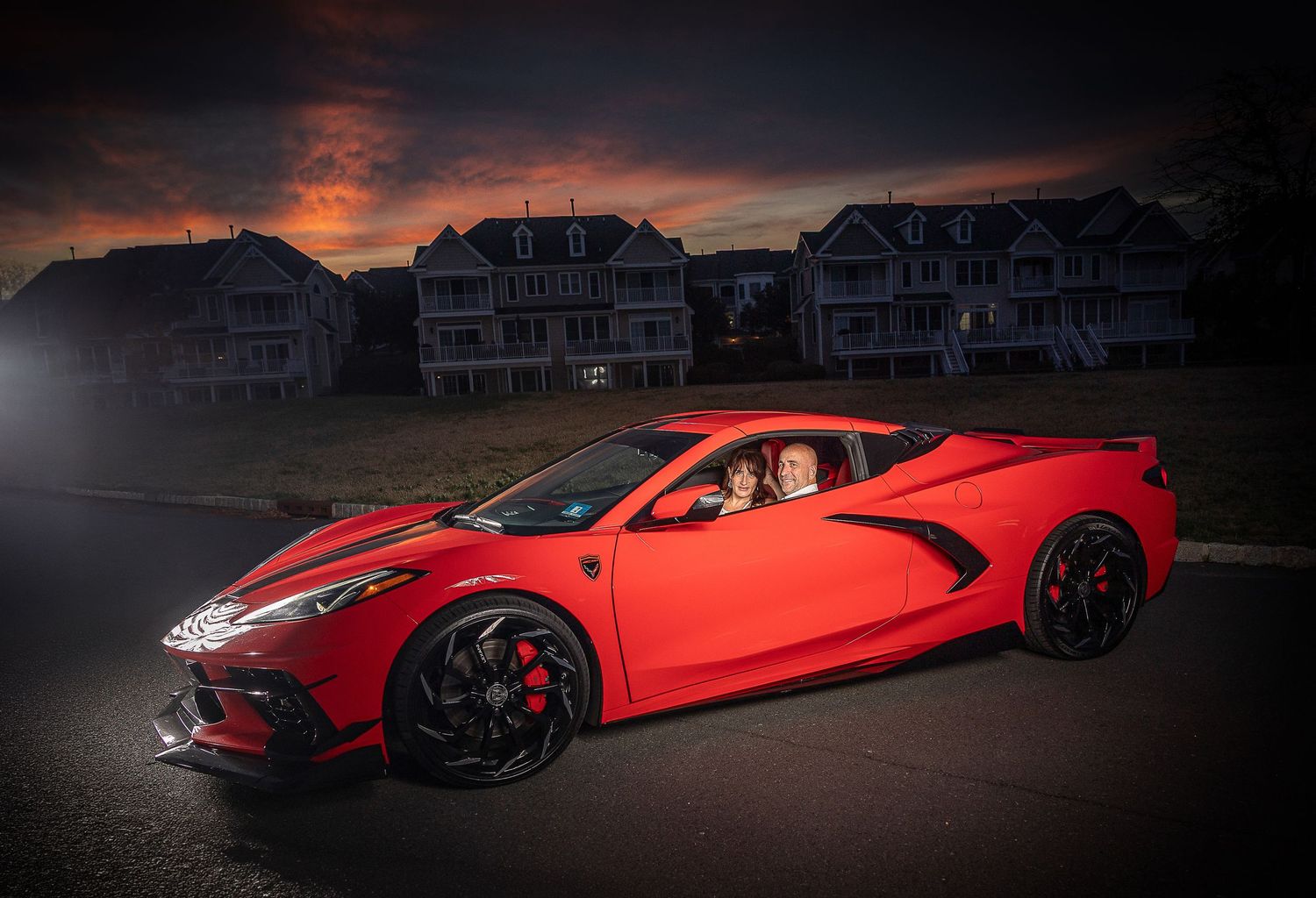 Red mid-engine Corvette C8 sports car parked at dusk with dramatic sky and suburban homes in background.