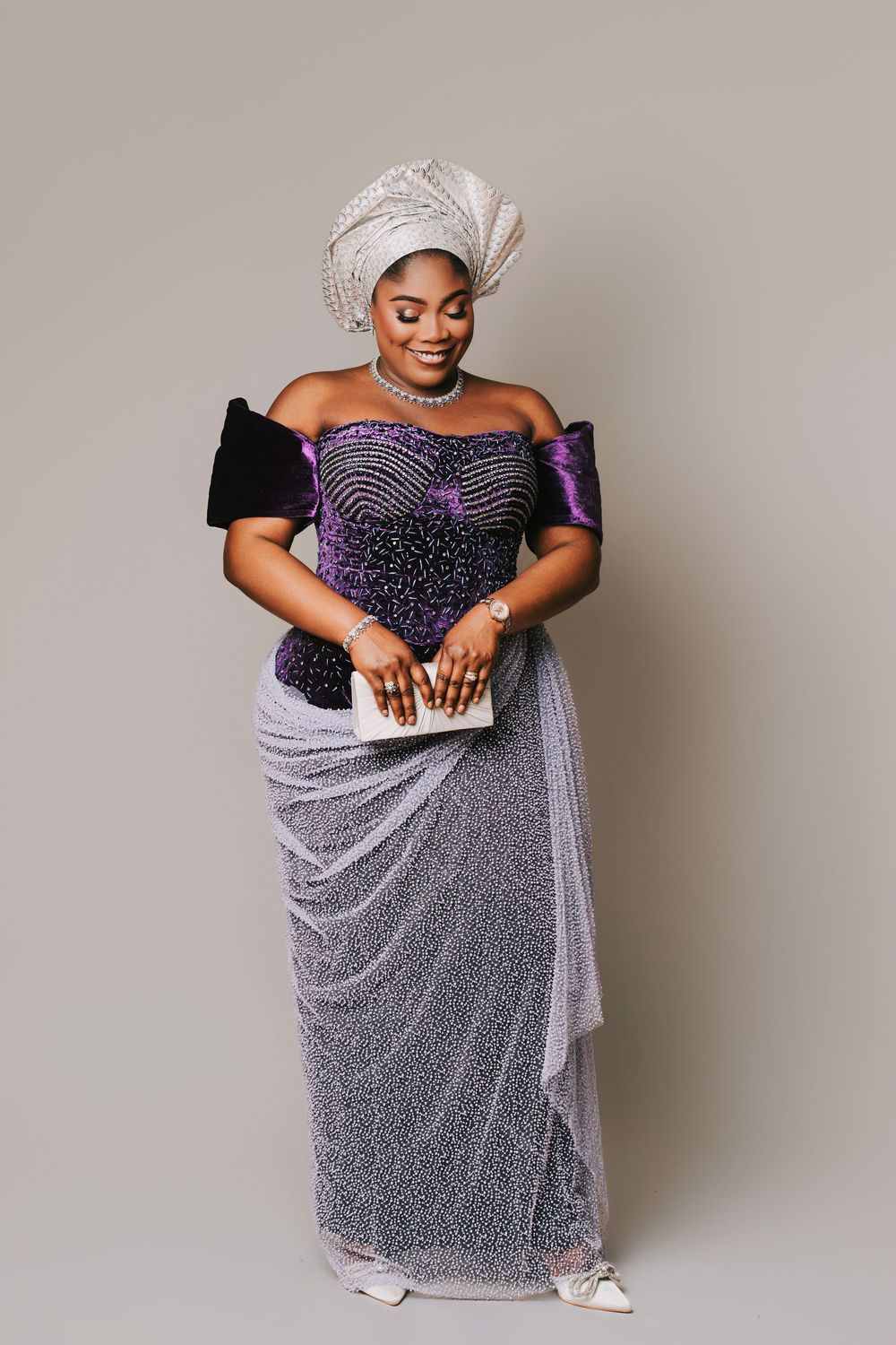 Woman in elegant purple beaded off-shoulder gown with silver head wrap and clutch purse in traditional African attire.