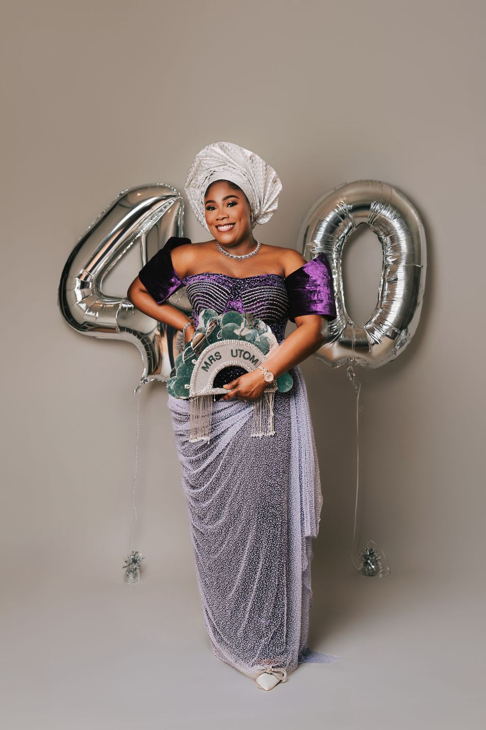 Woman in traditional Nigerian attire celebrates 40th birthday with silver balloons, wearing a gele headwrap and holding a decorative fan.