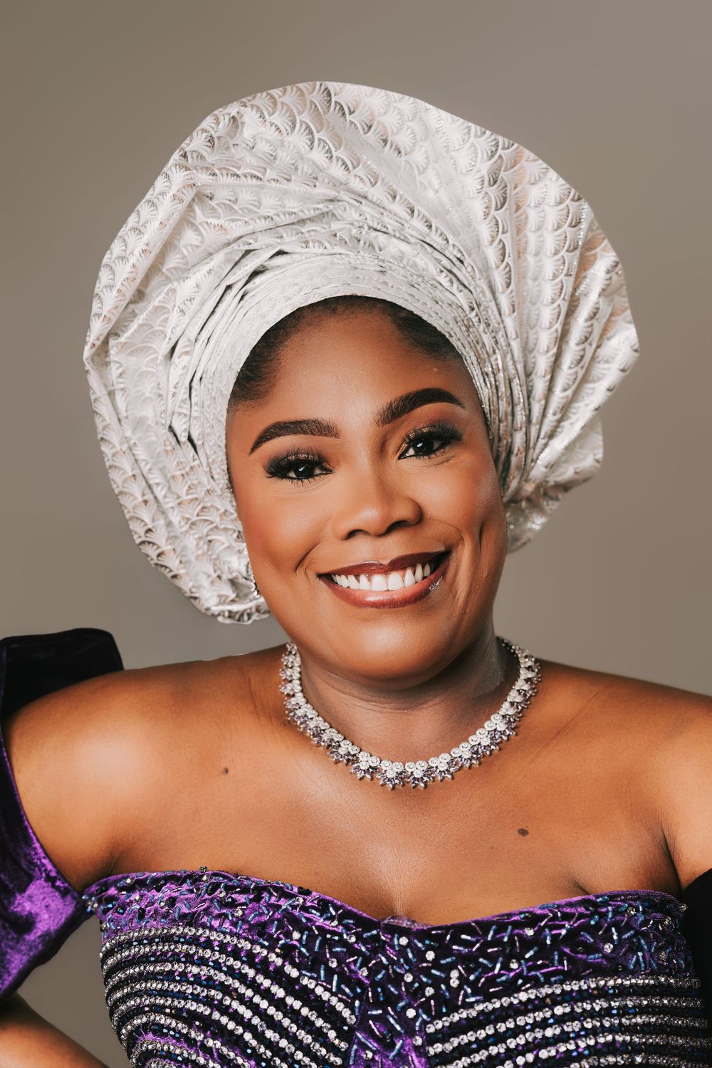 Smiling woman wearing a white gele headwrap, diamond necklace, and purple beaded outfit in a portrait photo.