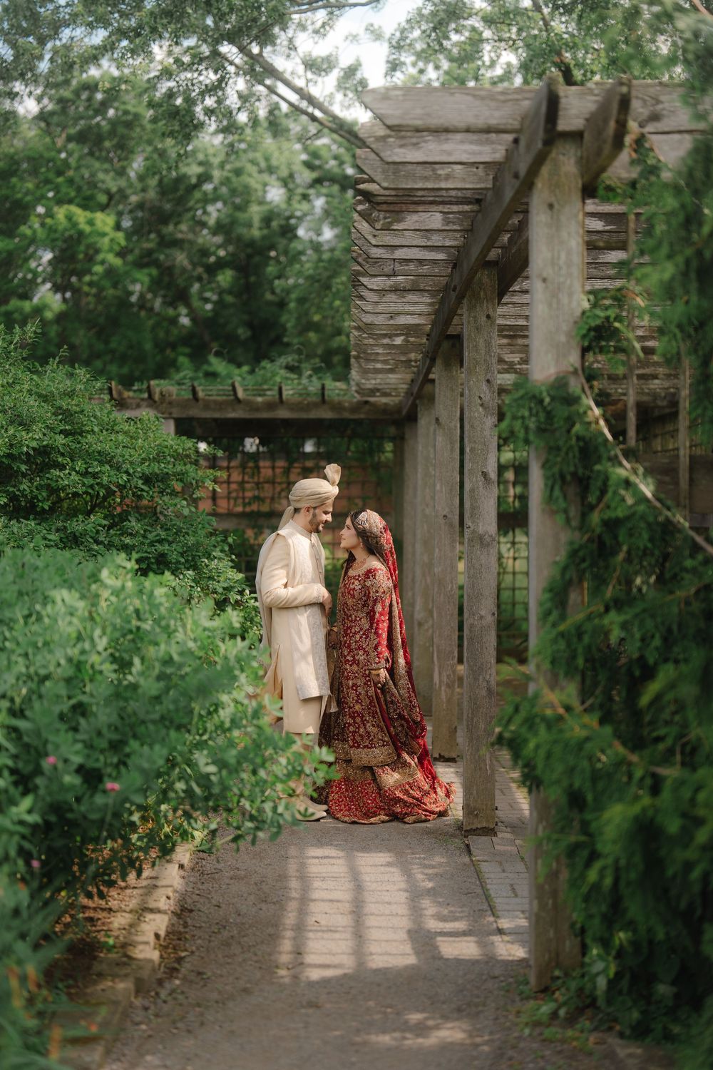 Indian bride and groom in traditional attire standing under a wooden pergola surrounded by lush greenery.