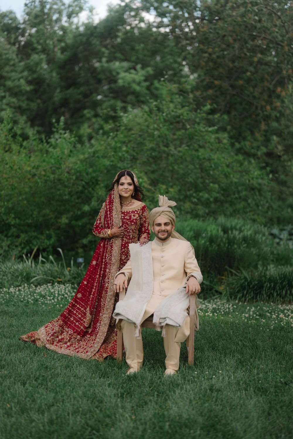 South Asian bride in red lehenga stands beside groom in cream sherwani seated outdoors in lush garden wedding portrait.