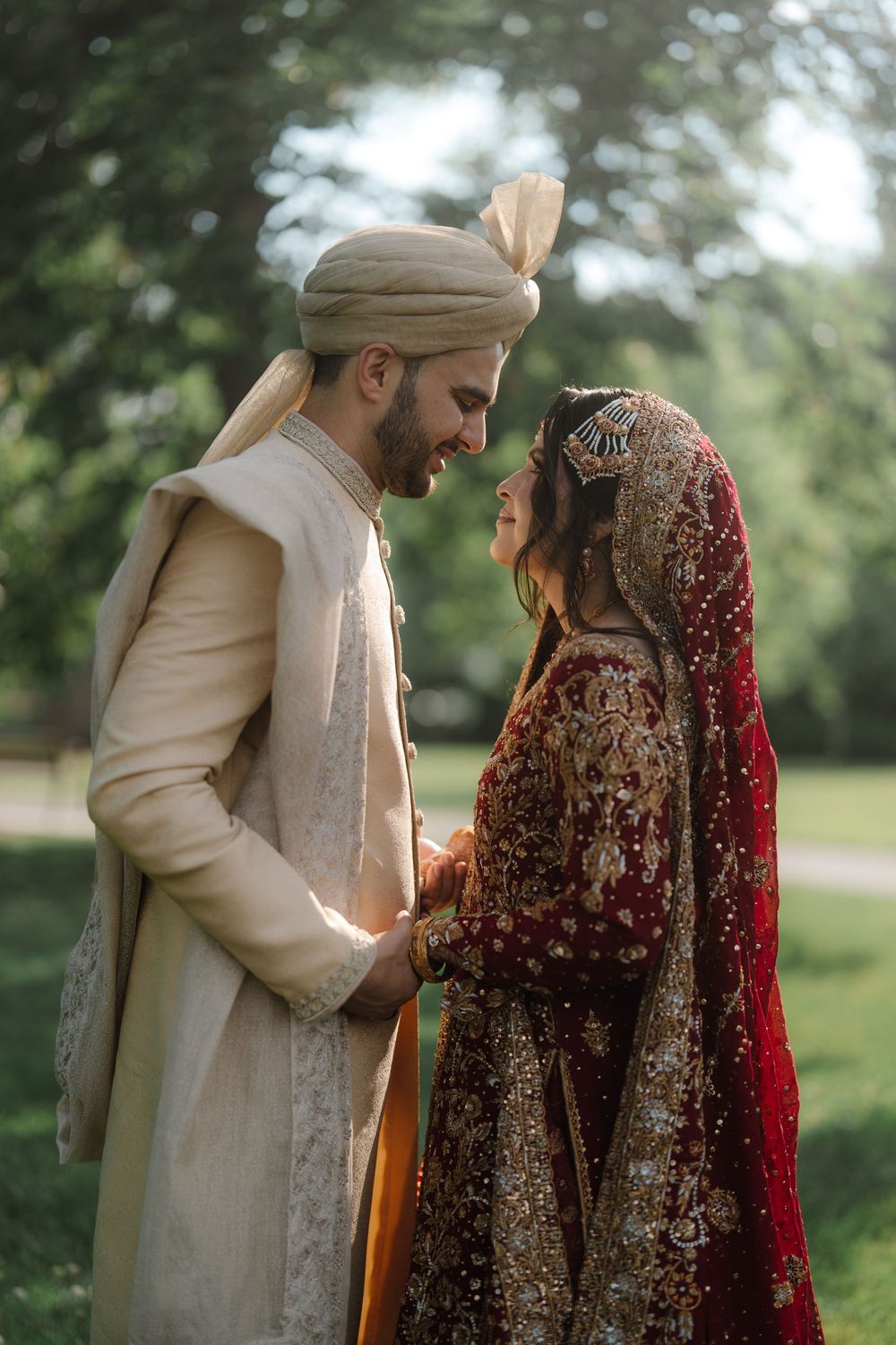 South Asian bride and groom in traditional wedding attire holding hands outdoors, wearing sherwani and red bridal lehenga.