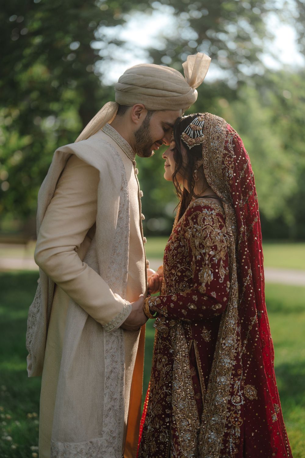 South Asian bride and groom in traditional wedding attire share an intimate moment outdoors, touching foreheads tenderly.