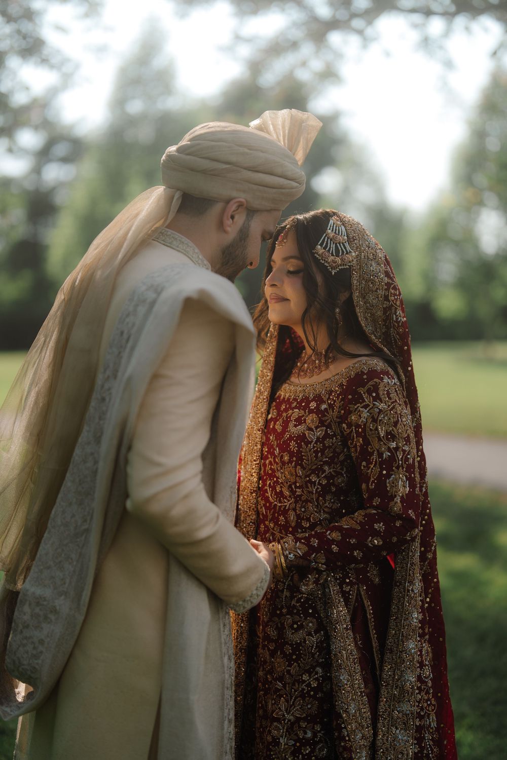 South Asian bride in red lehenga and groom in cream sherwani share an intimate moment in a lush garden setting.