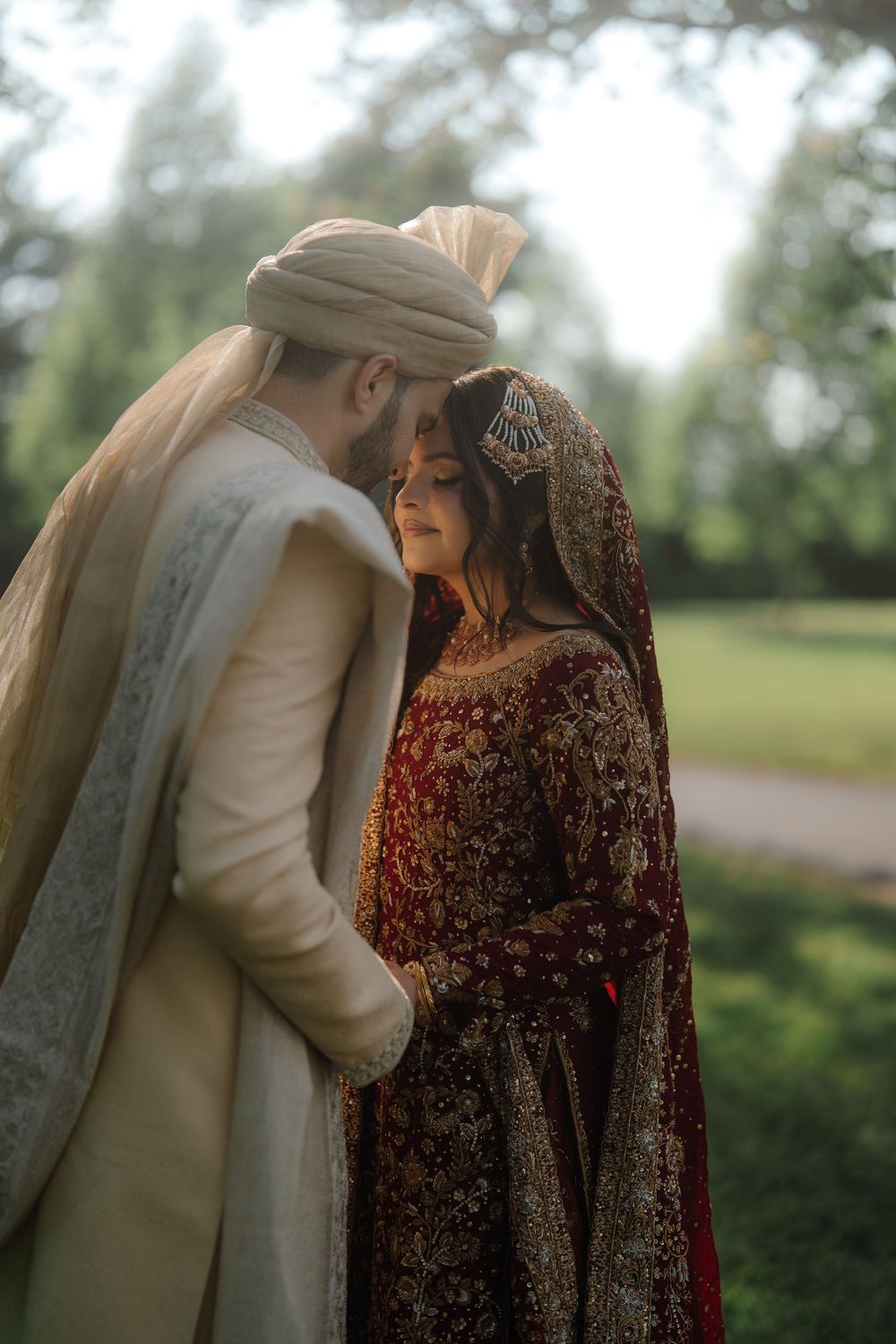 South Asian bride and groom in traditional wedding attire share a romantic moment outdoors, surrounded by lush greenery.