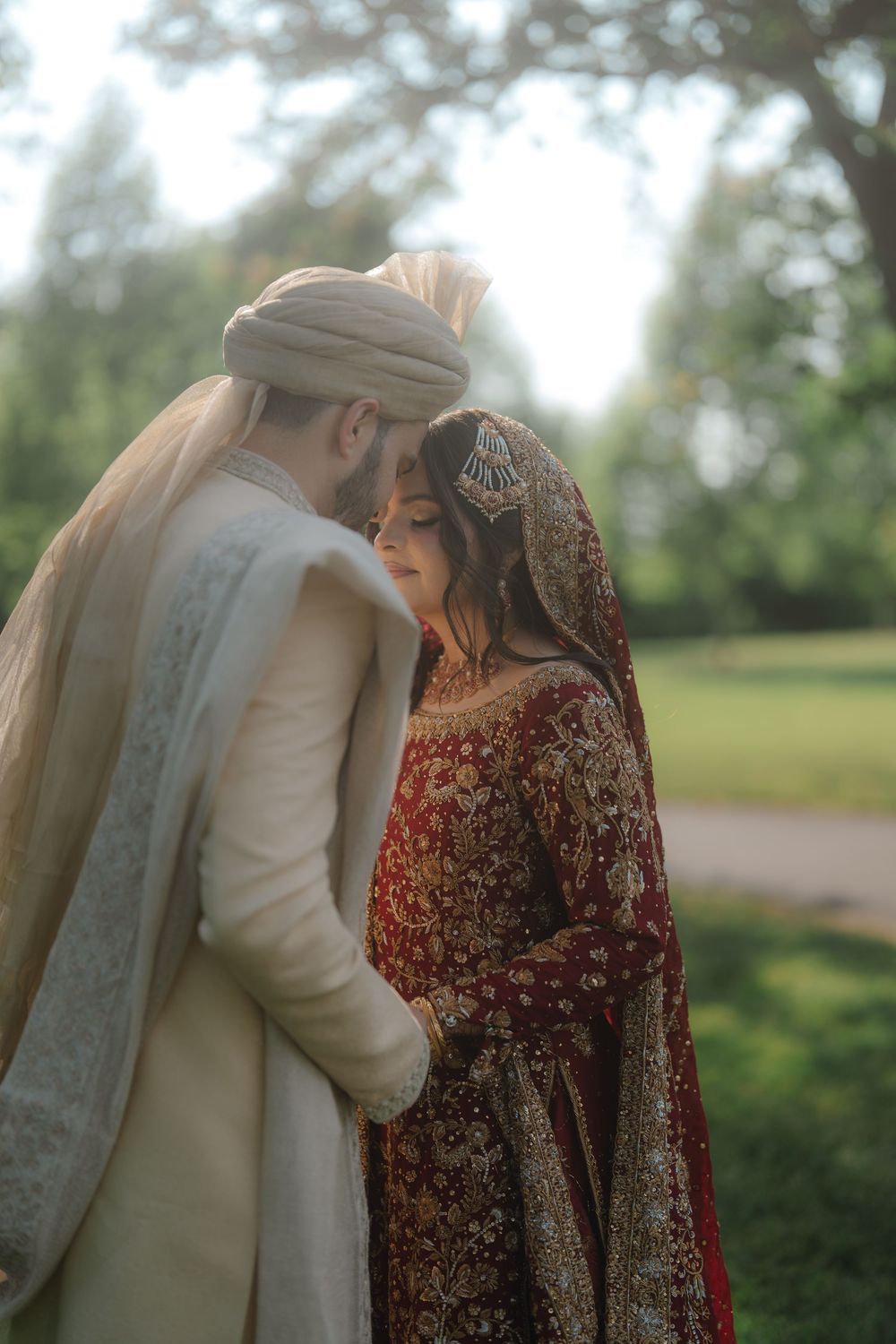 A couple in traditional South Asian wedding attire share an intimate moment outdoors, bride in red lehenga and groom in cream sherwani.